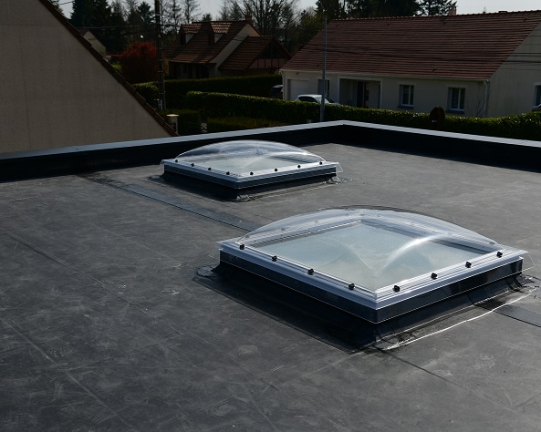 Toit plat sombre avec deux dômes de lumière transparents et encadrements argentés. Maisons et arbres en arrière-plan sous un ciel clair.