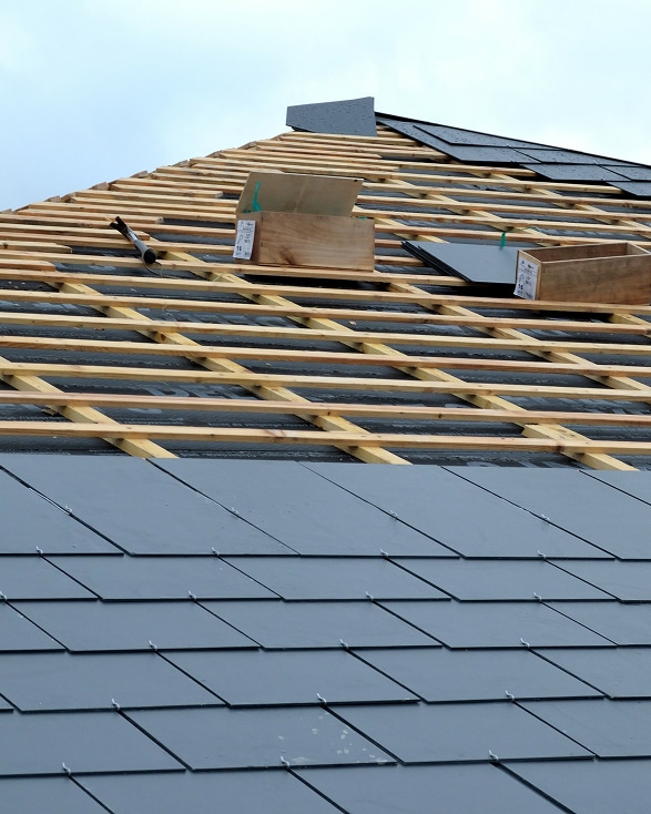 Toiture en construction : ardoises grises posées, lattes de bois apparentes, avec matériaux et outils sur les chevrons.