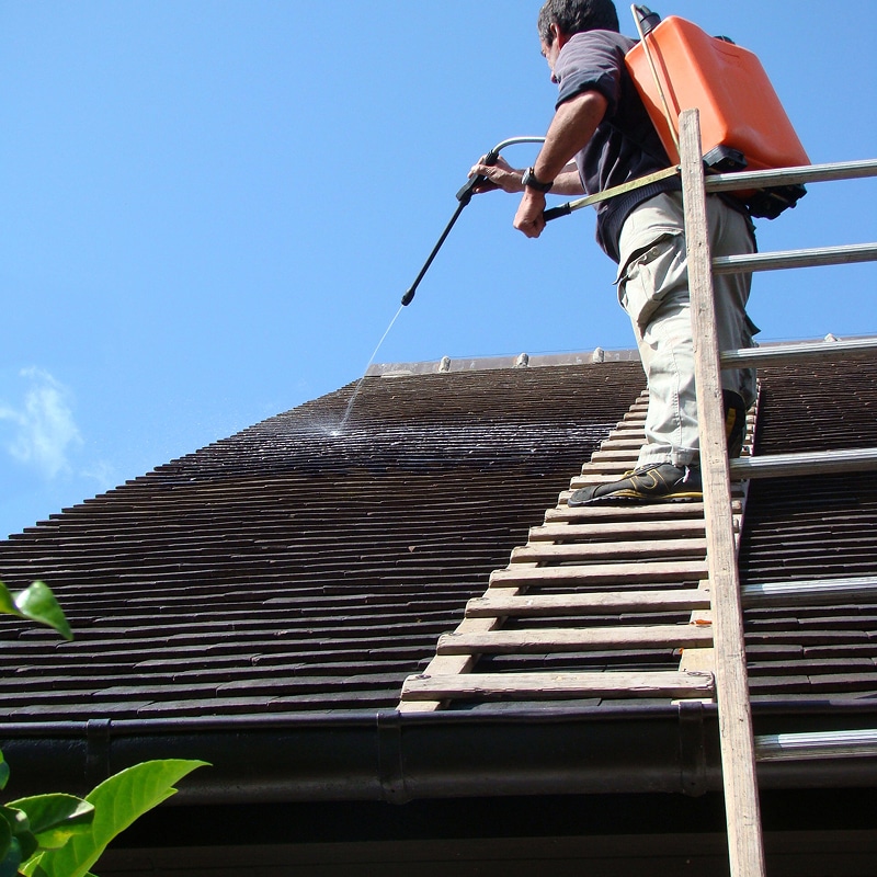 Un homme sur une échelle de toit pulvérise un produit pour nettoyer les tuiles d'un toit résidentiel sous un ciel bleu clair.
