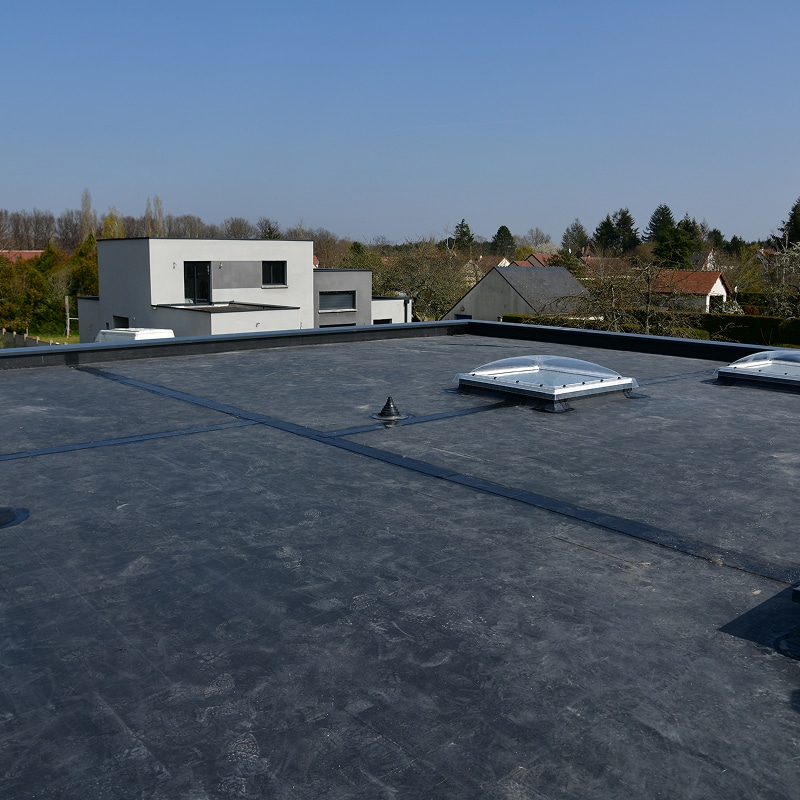 Toit plat gris foncé avec membrane d'étanchéité, deux puits de lumière dôme, un évent, et empreintes de pas. Maison moderne en arrière-plan sous ciel bleu.