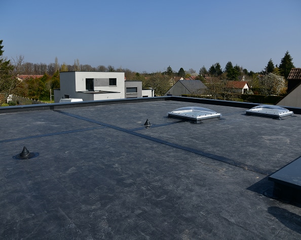 Toit plat gris foncé avec deux puits de lumière en dôme, un rectangulaire et des aérations. Maison moderne en arrière-plan, ciel clair.
