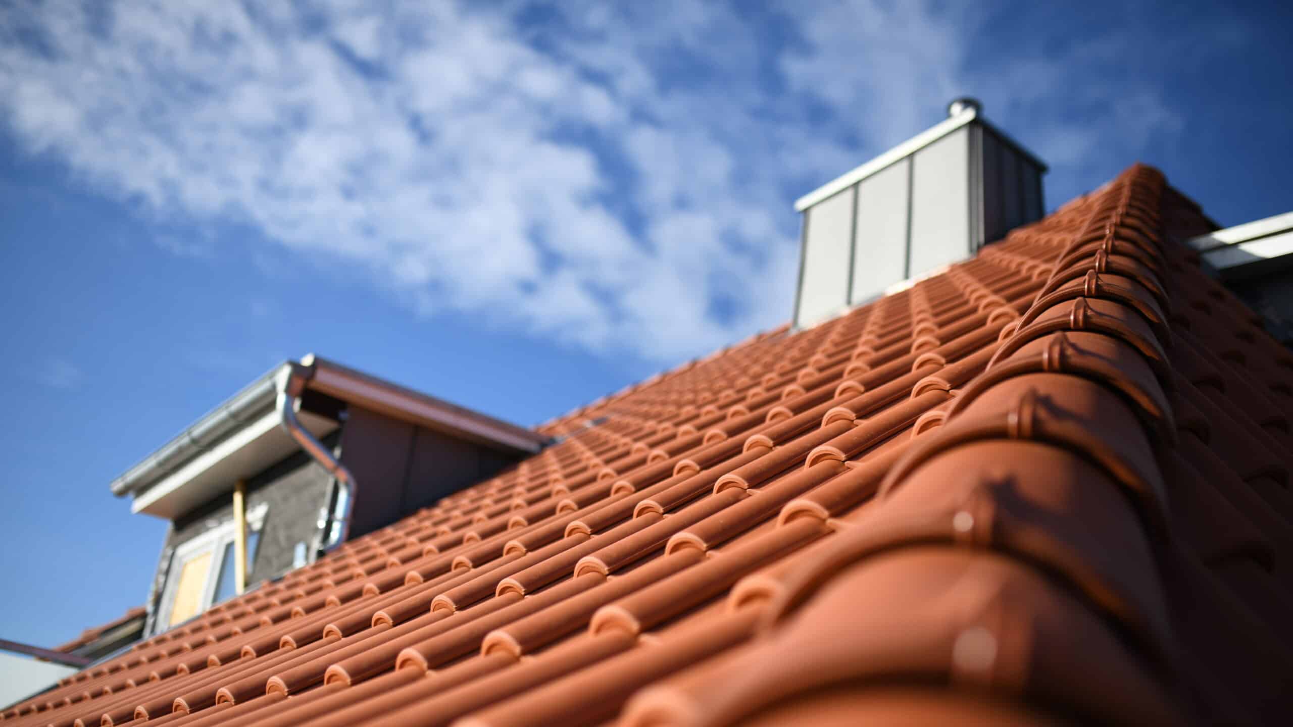 Toit résidentiel en tuiles de terre cuite rouges sous un ciel bleu parsemé de nuages, avec cheminée et gouttière visibles.