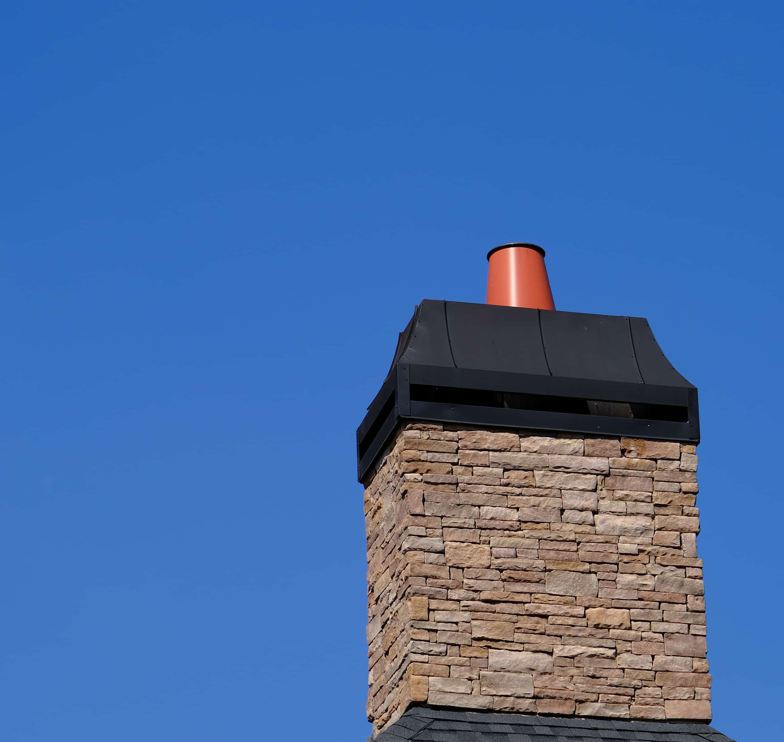 Cheminée moderne en pierres naturelles, coiffe métallique noire et conduit orange vif. Ciel bleu éclatant en arrière-plan.