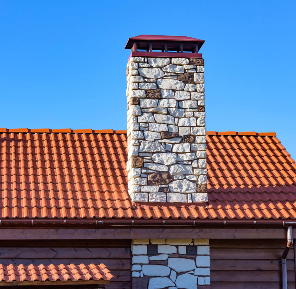 Cheminée en pierre décorative et toit de tuiles terracotta sur une maison en bois, avec gouttière et ciel bleu.