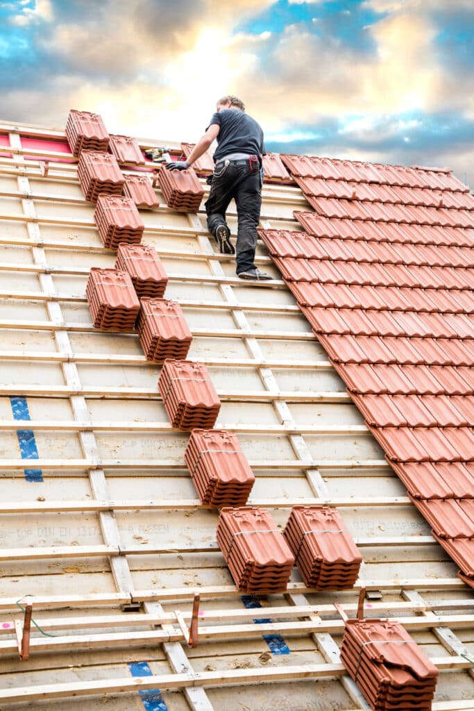 Un couvreur pose des tuiles en terre cuite sur un toit en construction. Des piles de tuiles sont alignées sur les lattes en bois sous un ciel nuageux.