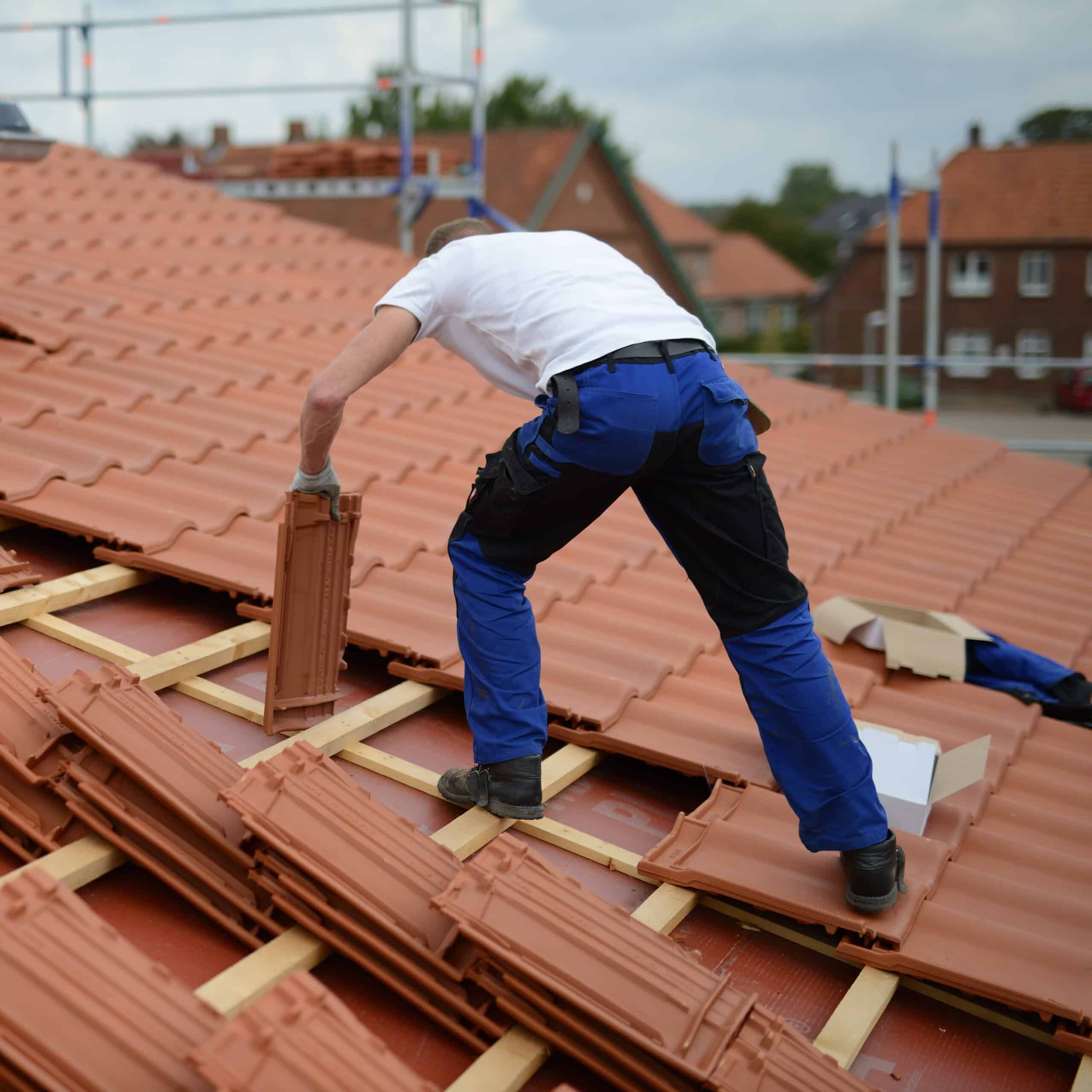 Couvreur avec gants installant des tuiles en terre cuite sur un toit en construction, avec des lattes en bois et d'autres toits au loin.