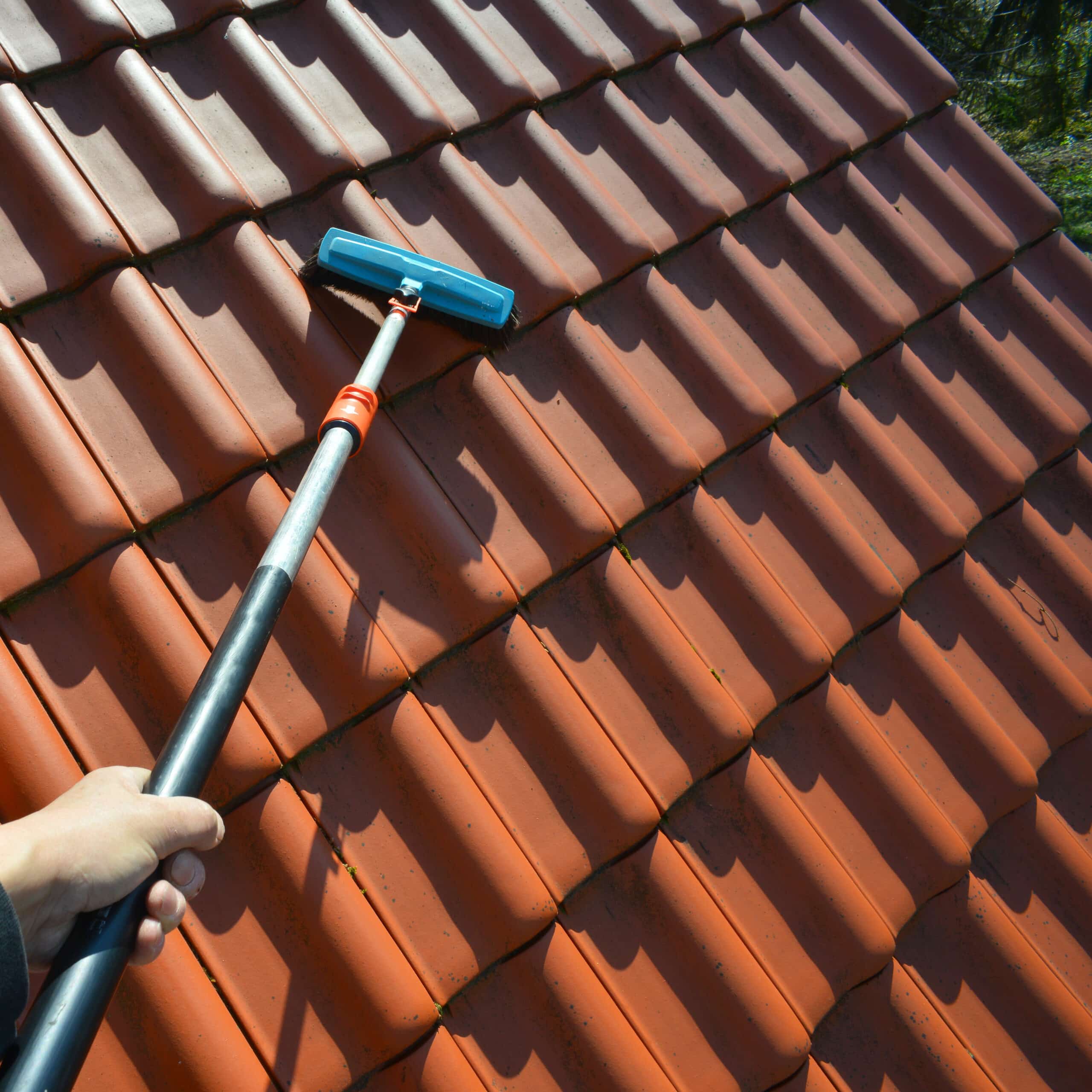 Nettoyage de toit en tuiles terracotta. Une main utilise une brosse sur perche pour éliminer saleté et mousse sous un soleil intense.