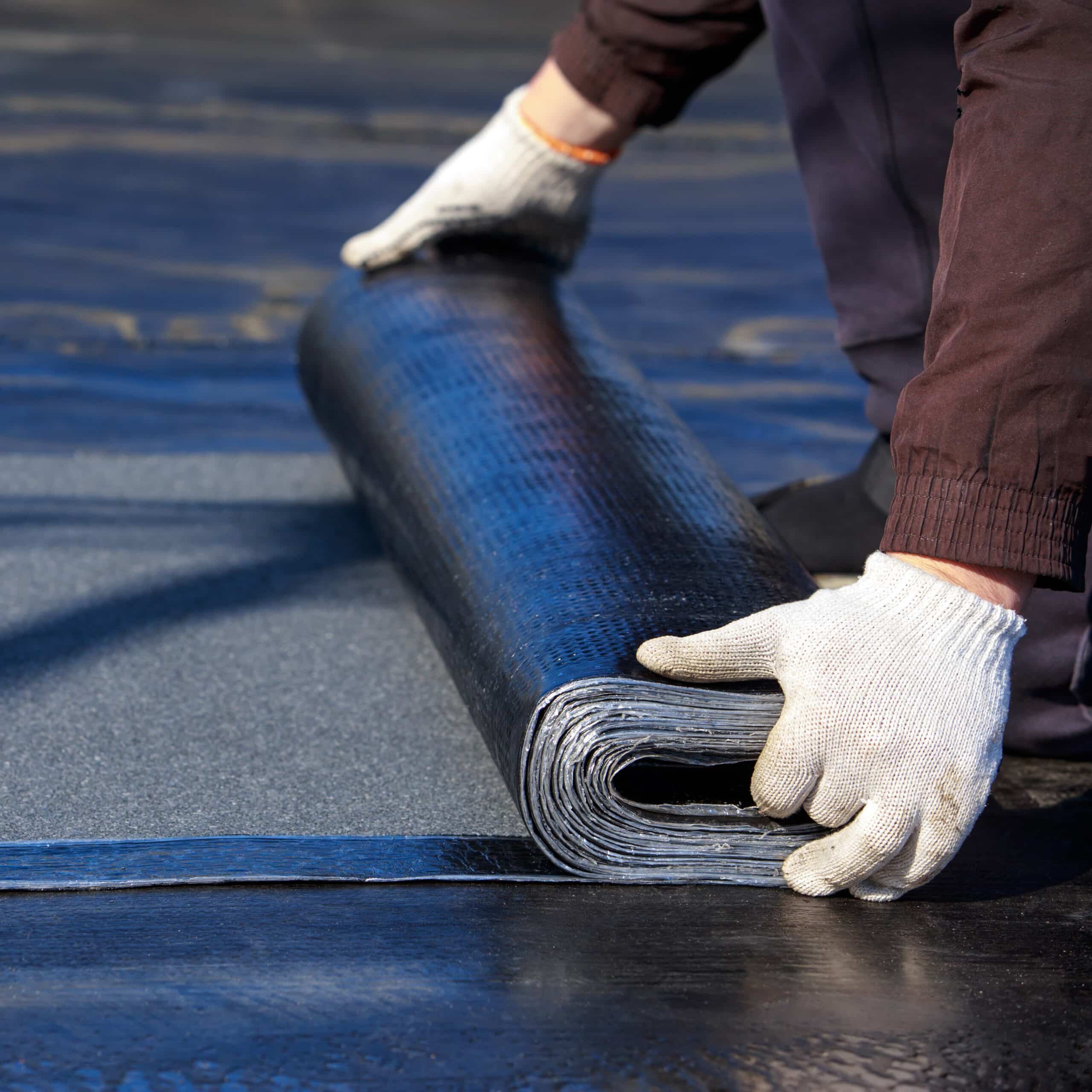 Ouvrier en gants déroulant un rouleau de membrane d'étanchéité bitumineuse noire sur une toiture plate pour la pose.
