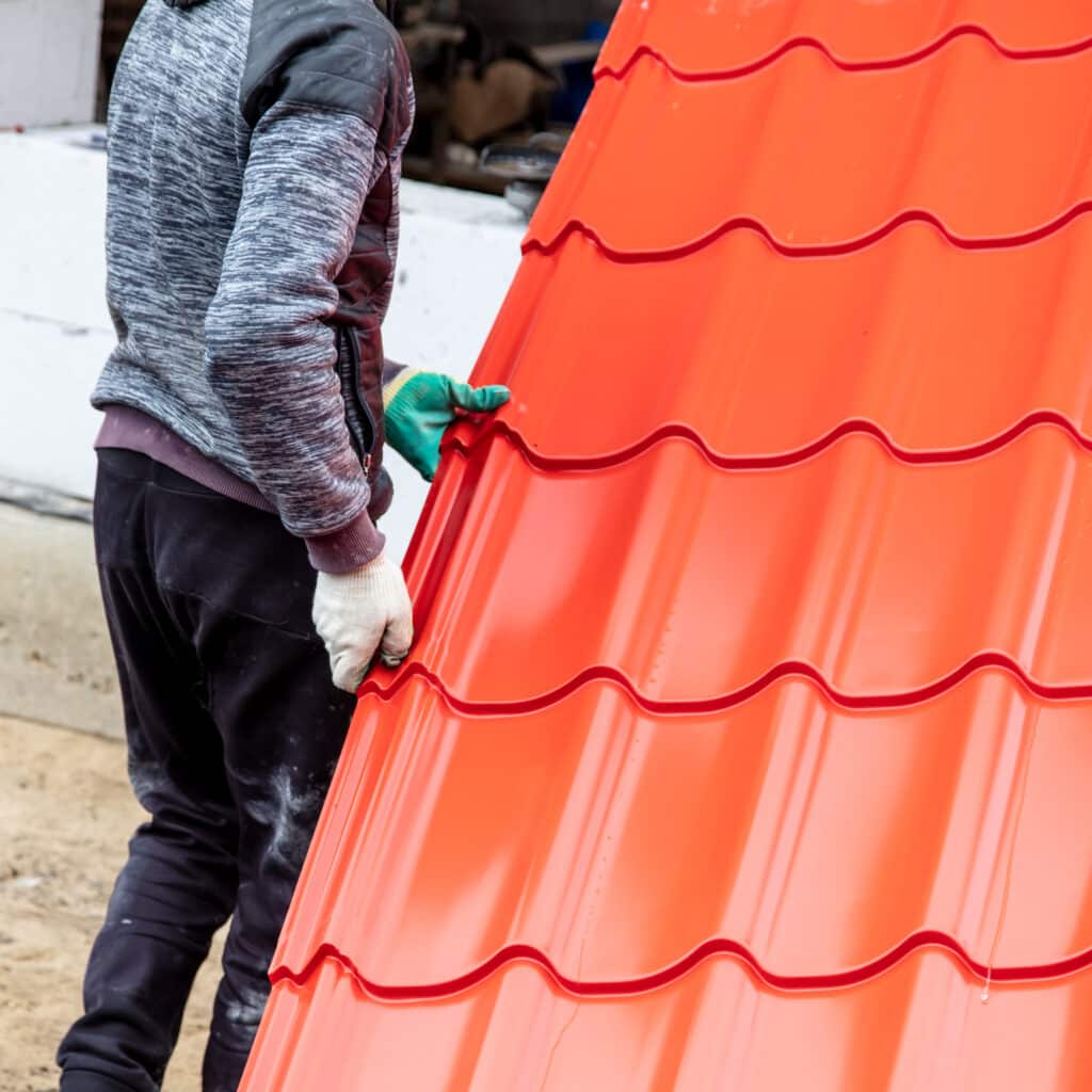Ouvrier en gants manipulant une tôle de toiture métallique rouge brillante. Le panneau ondulé est prêt pour la pose sur un chantier.